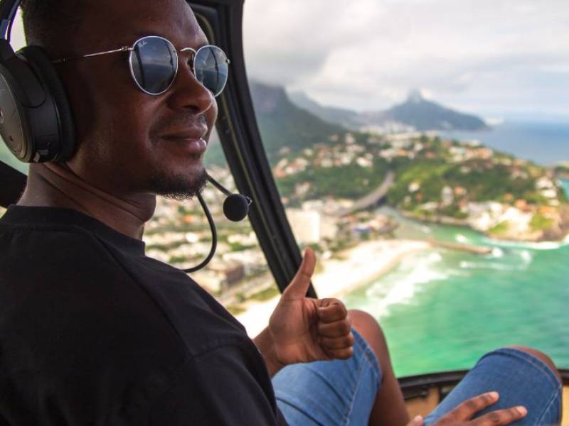 Homem fazendo joia com a mão e paisagem do Rio de Janeiro ao fundo durante voo panorâmico de helicóptero.